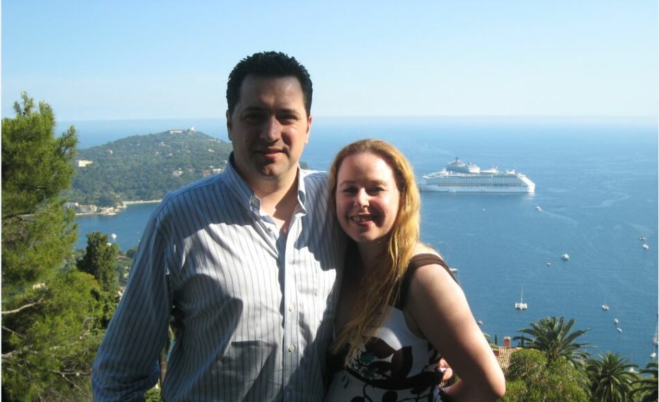 A man and woman travel around the world, striking a pose in front of a cruise ship.