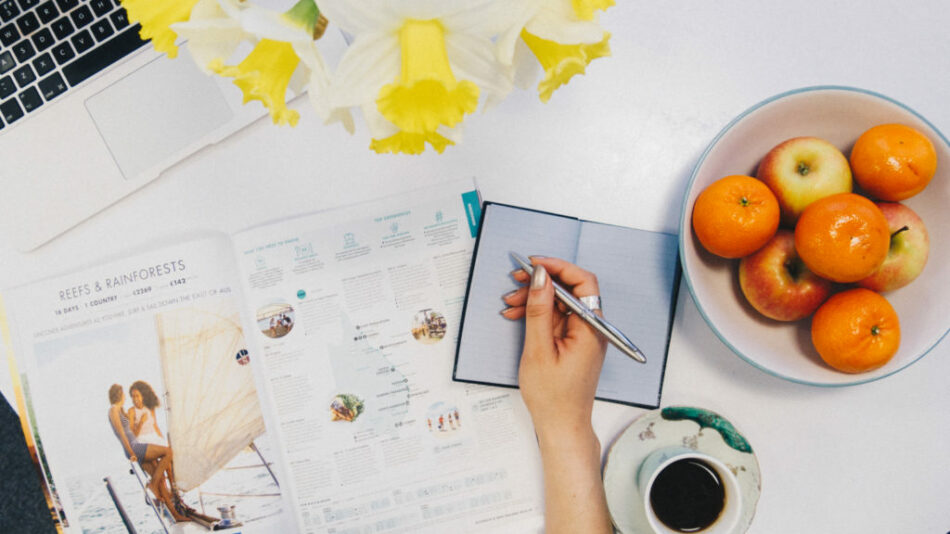 A woman is writing on a notebook while sitting at a desk with oranges and a laptop.