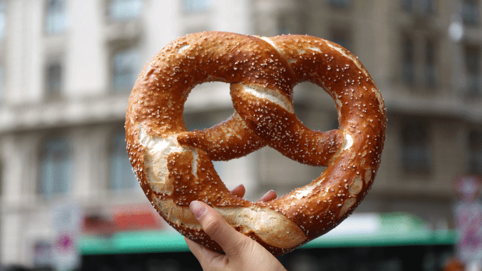 A person holding a German pretzel in front of a building.