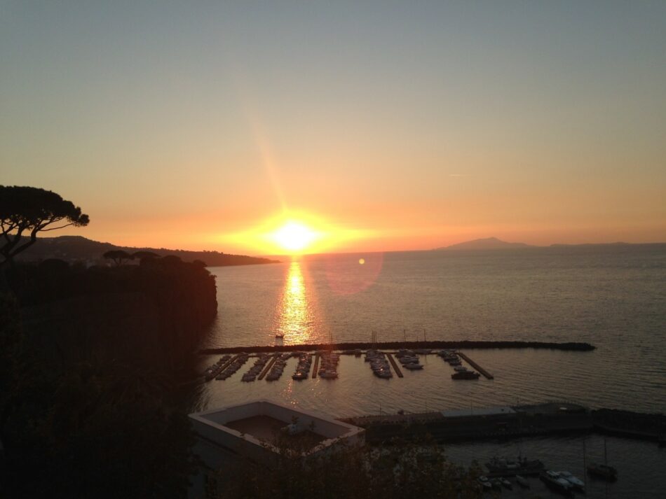 Travel to Italy and watch the sun setting over a charming harbor with boats docked.