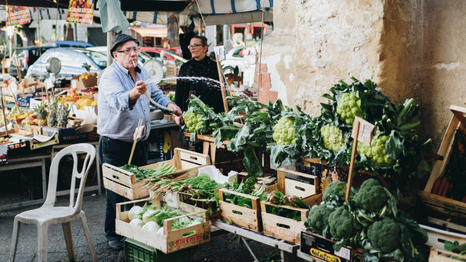 One of the popular things to do in Sicily is to visit the vibrant local markets. In a bustling market, a man proudly stands behind his colorful vegetable stall, offering a variety of fresh