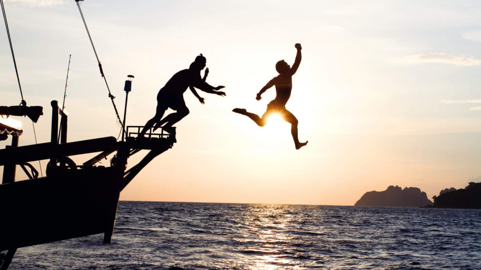 Two people jumping off a boat at sunset while capturing the moment in the best travel books.