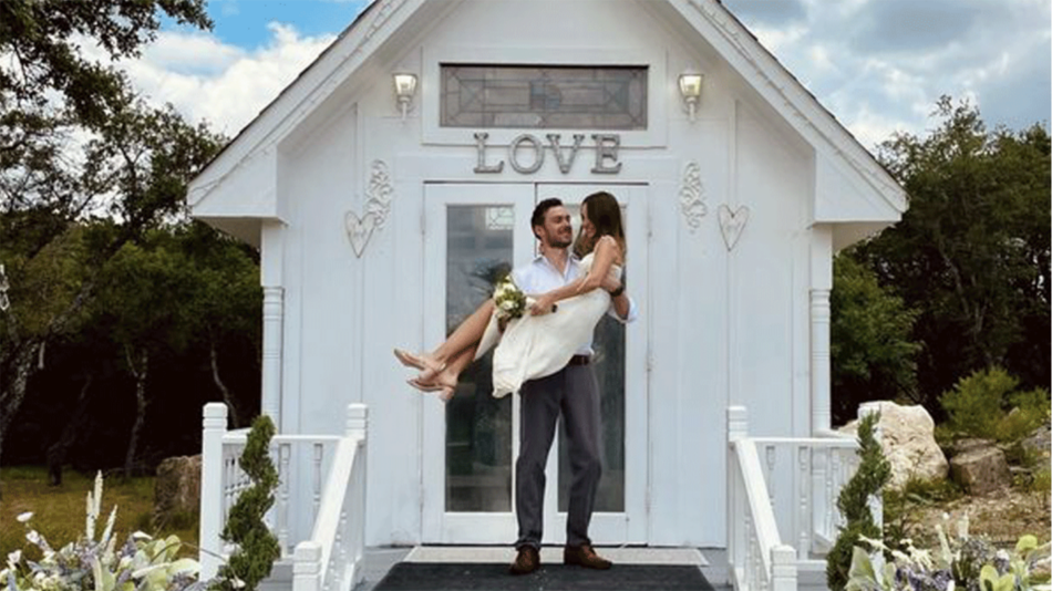 A real love story unfolds as a bride and groom stand in front of a small white house.