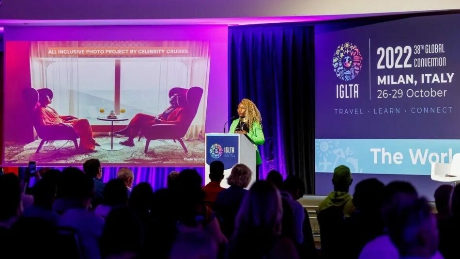 A woman is speaking at a conference about LGBTQIA+ travel in front of a large screen.