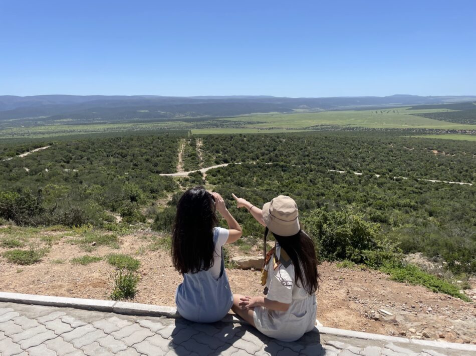 Two women, one being a safari ranger, sitting on the edge of a hill overlooking a valley.