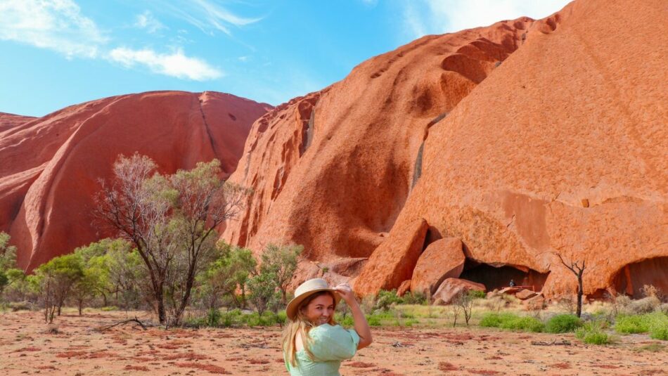 Female solo traveller in her early twenties, clothed in green dress and Australian wide-brimmed hat, stands in front of Uluru.